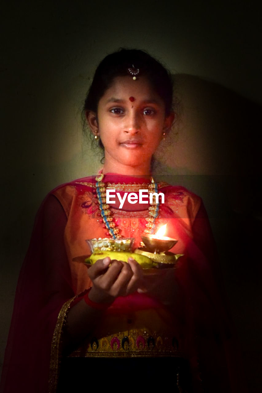 one person, portrait, looking at camera, women, indoors, adult, clothing, traditional clothing, front view, jewelry, red, waist up, necklace, emotion, studio shot, person, smiling, celebration, young adult, tradition, bangle, religion, female, bracelet, sari, holding, event, standing, child, fashion accessory, temple, belief