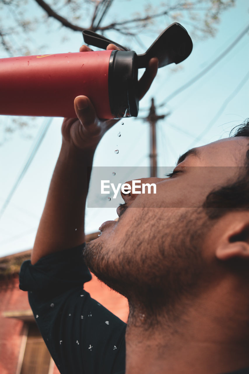 Close-up of young man pouring water on face against sky