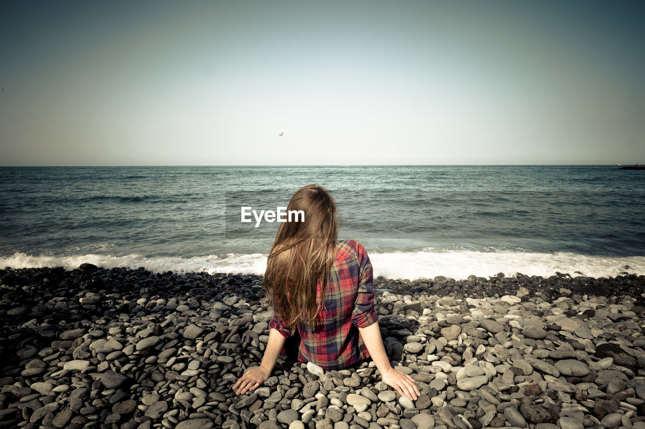 Rear view of woman sitting on rock at beach against clear sky