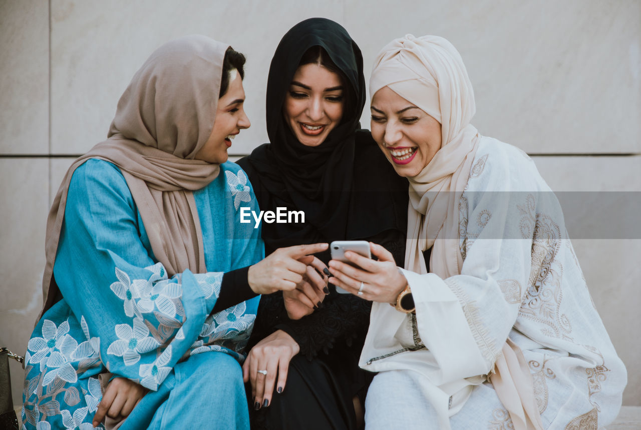 Cheerful friends in hijab looking at phone sitting against wall