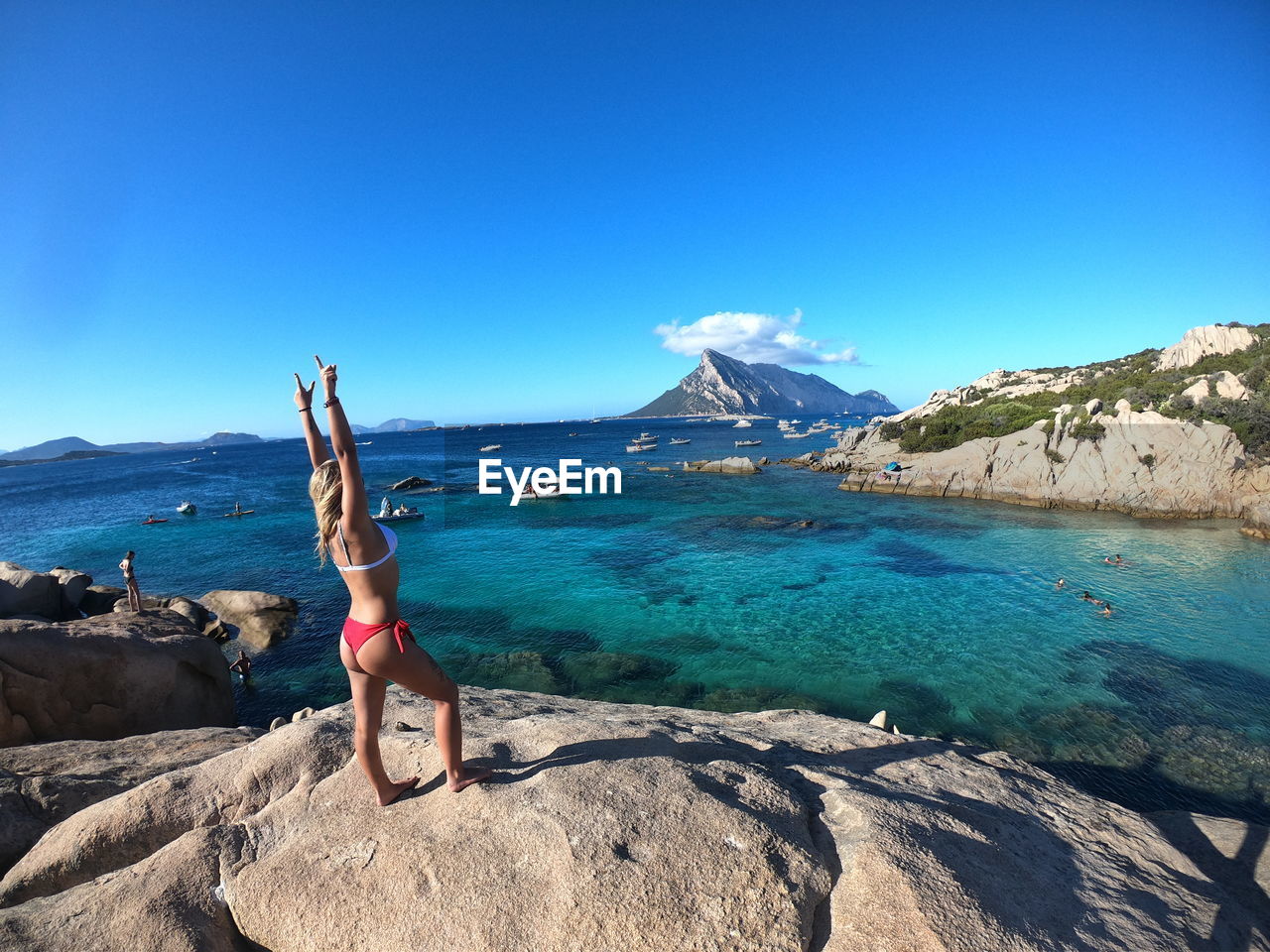 Woman in bikini standing on rock by sea against blue sky