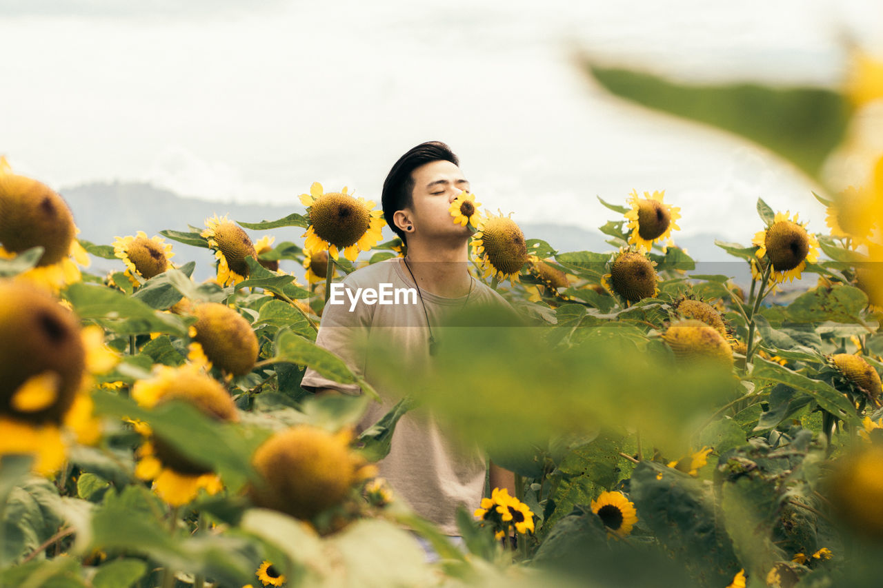 Young man with closed eyes standing amidst sunflowers against sky