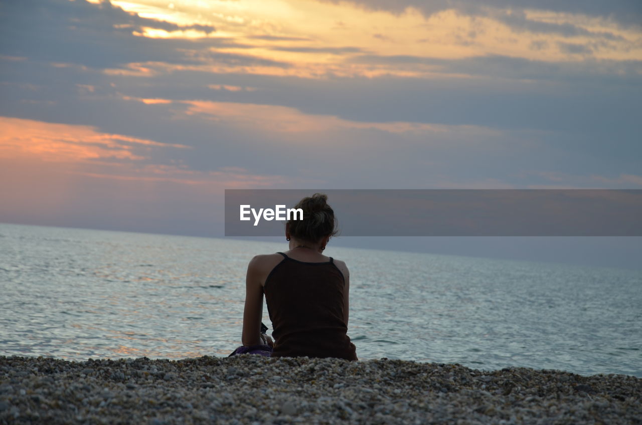 Wife sitting on shore against sky during sunset