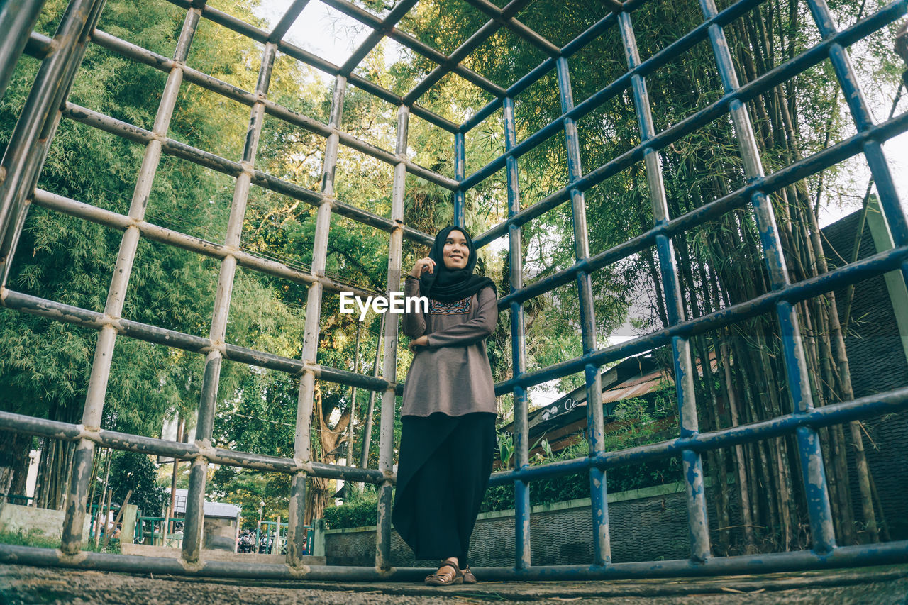 Low angle view of smiling woman leaning on metal grate against trees at park