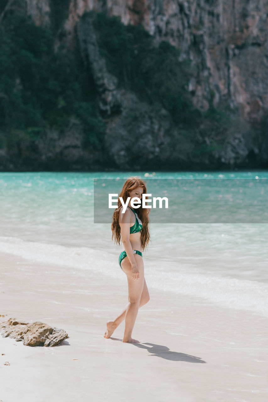 Full body side view of fit female in swimwear standing on sandy coast near azure sea against mountain in thailand