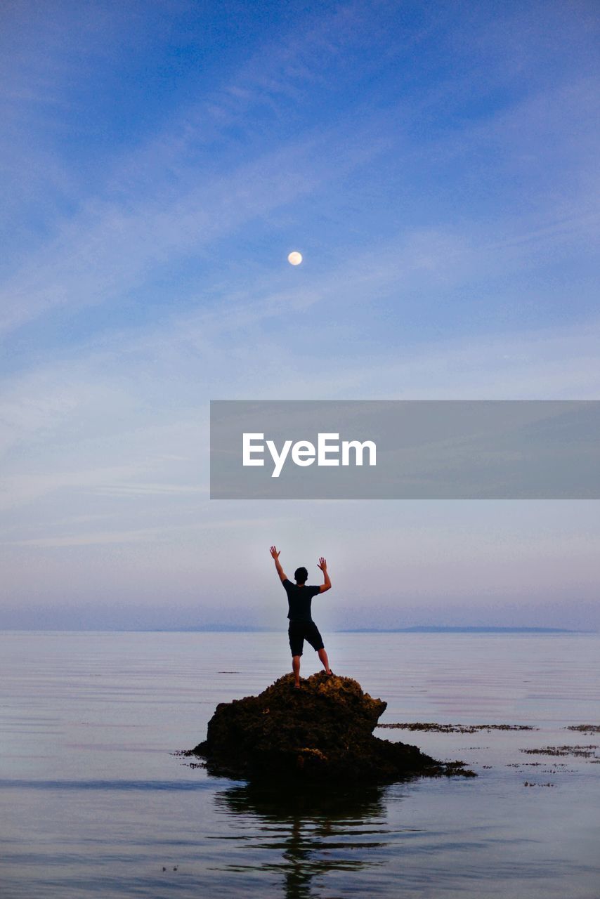 Rear view of man standing on rock amidst sea against sky