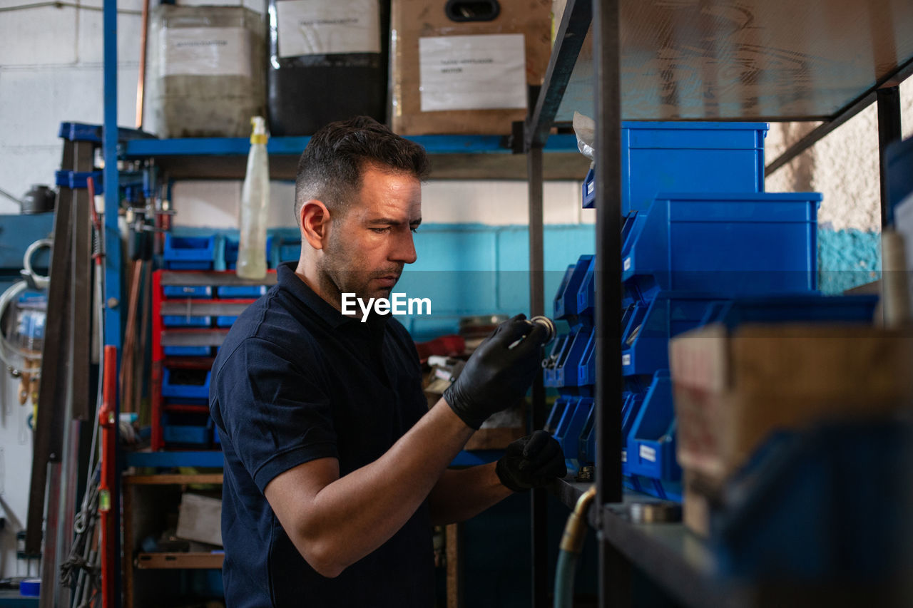 Positive adult male master in protective gloves picking metal detail from box while choosing tools for repair service in workshop