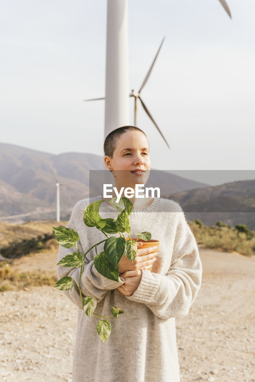 Shaved head woman holding potted plant at wind farm
