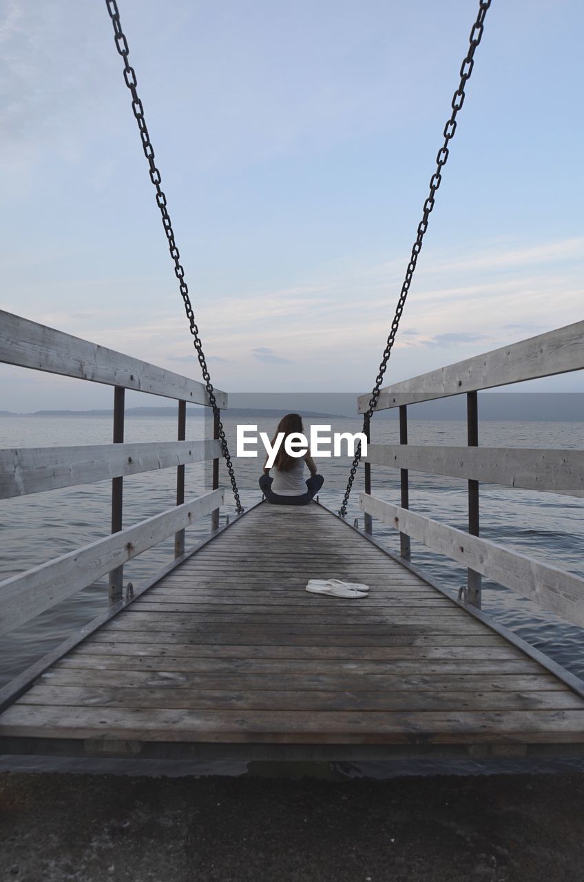 Rear view of woman sitting on jetty over sea against sky