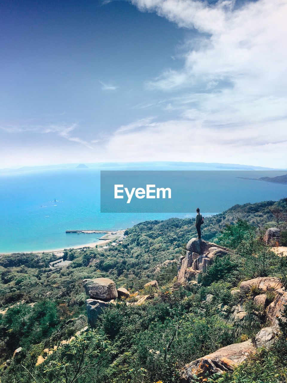 Aerial view of person standing on rock by sea against cloudy sky