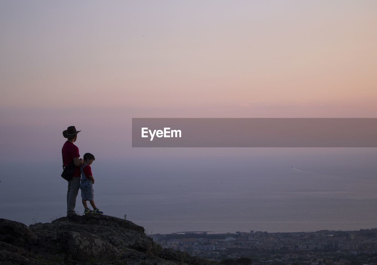 FULL LENGTH OF MAN STANDING ON ROCK AGAINST SKY AT SUNSET