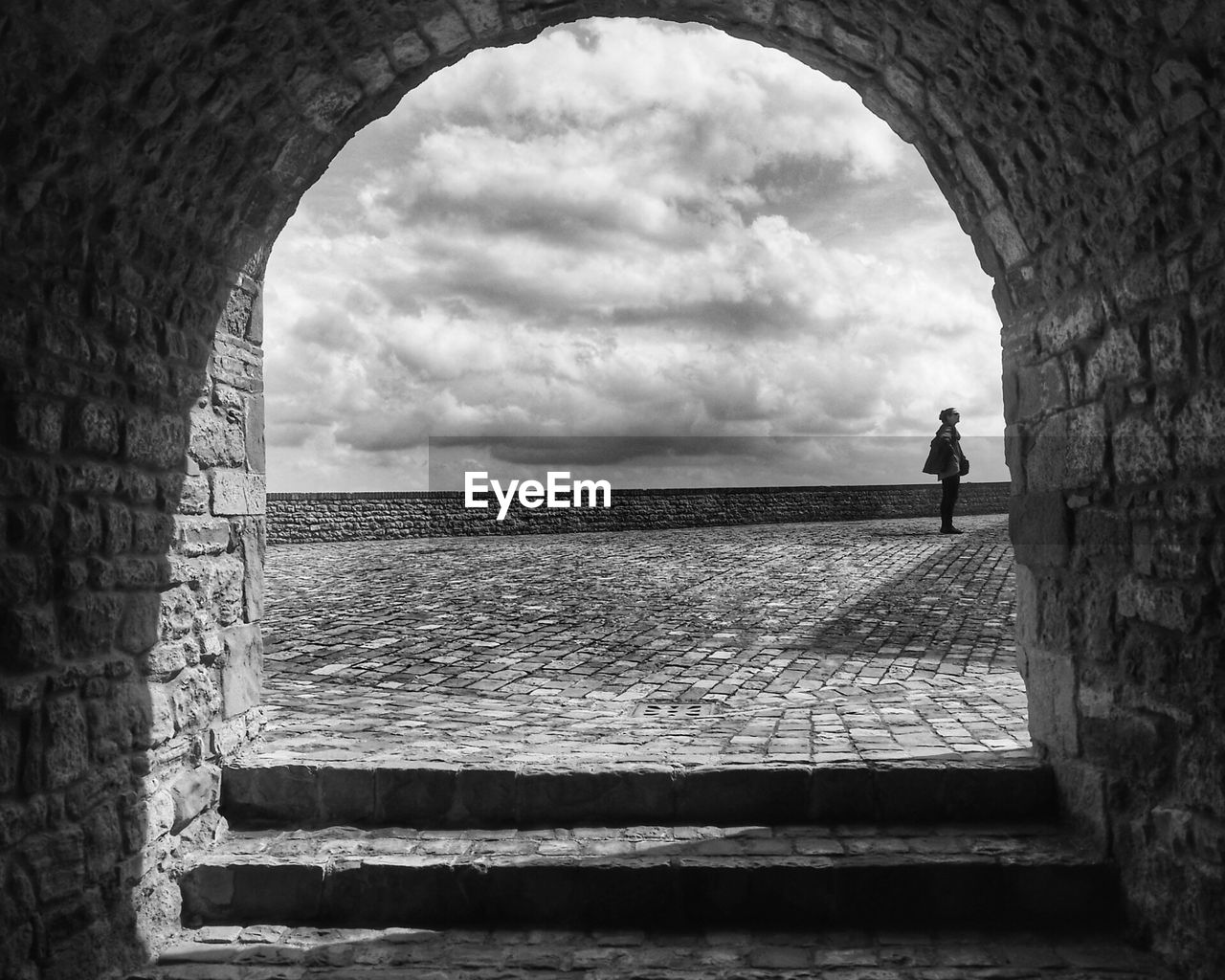 Woman standing on footpath against cloudy sky seen through arch