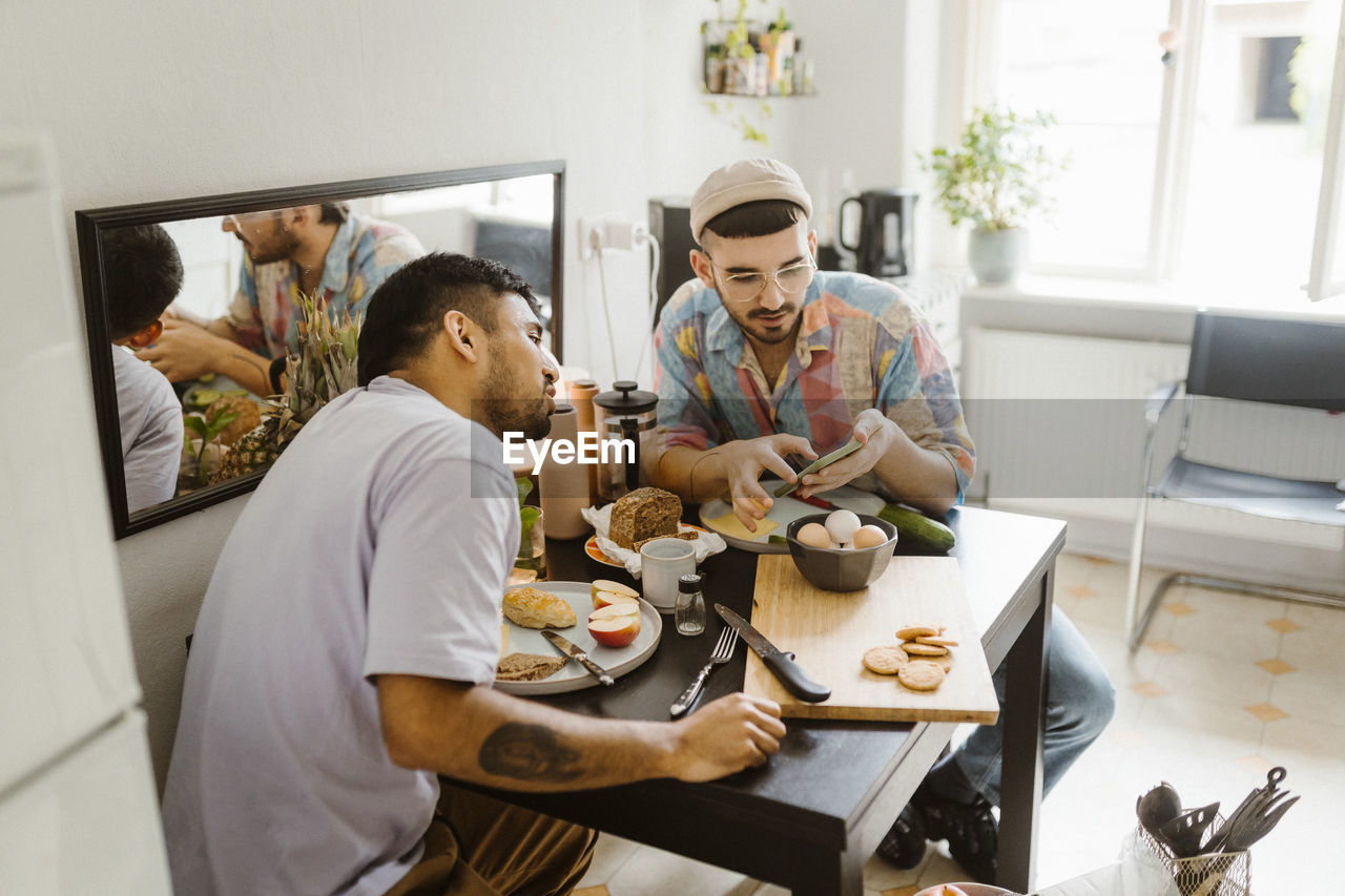 Gay couple sharing smart phone with each other while having food on dining table at home