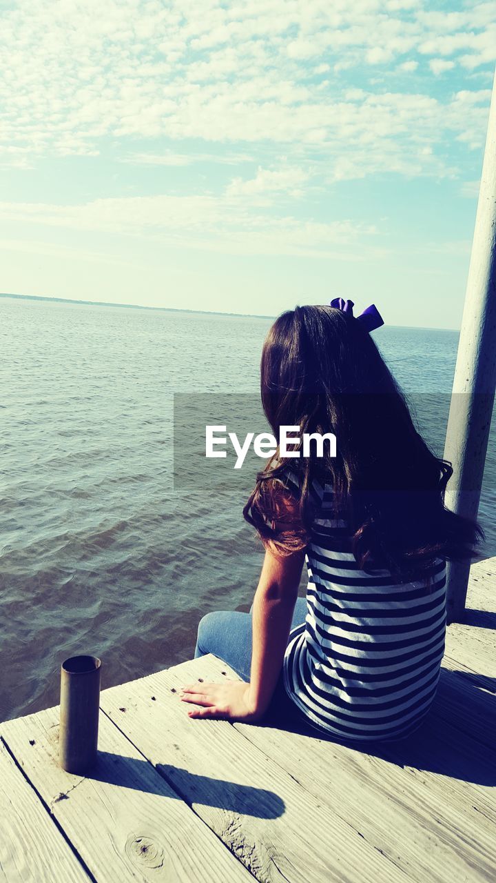 Rear view of girl sitting on boardwalk over sea against sky