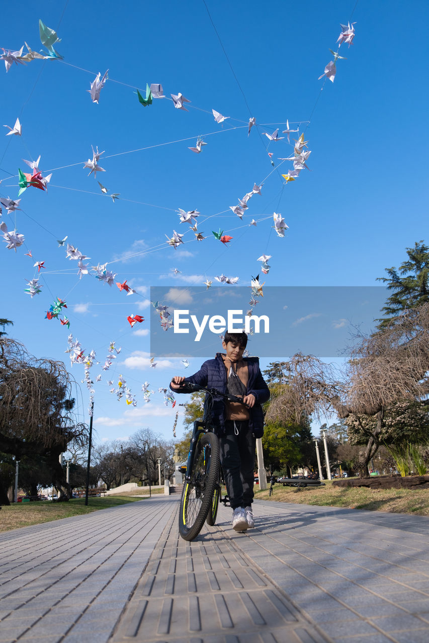 sky, transportation, one person, nature, full length, bicycle, motion, men, adult, blue, vehicle, wheel, day, mode of transportation, leisure activity, person, tree, clear sky, sports, on the move, outdoors, plant, city, sunny, sunlight, activity, bicycle wheel, cycling, extreme sports, casual clothing, headwear, front view, low angle view, land vehicle, lifestyles, helmet, road, architecture, travel, footpath, young adult, sports equipment, riding, flower