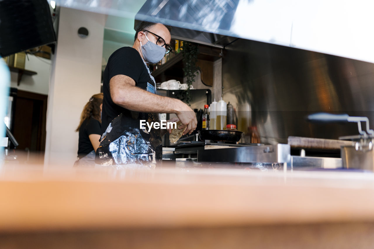Male barista wearing protective face mask while cooking in kitchen of coffee bar during covid-19