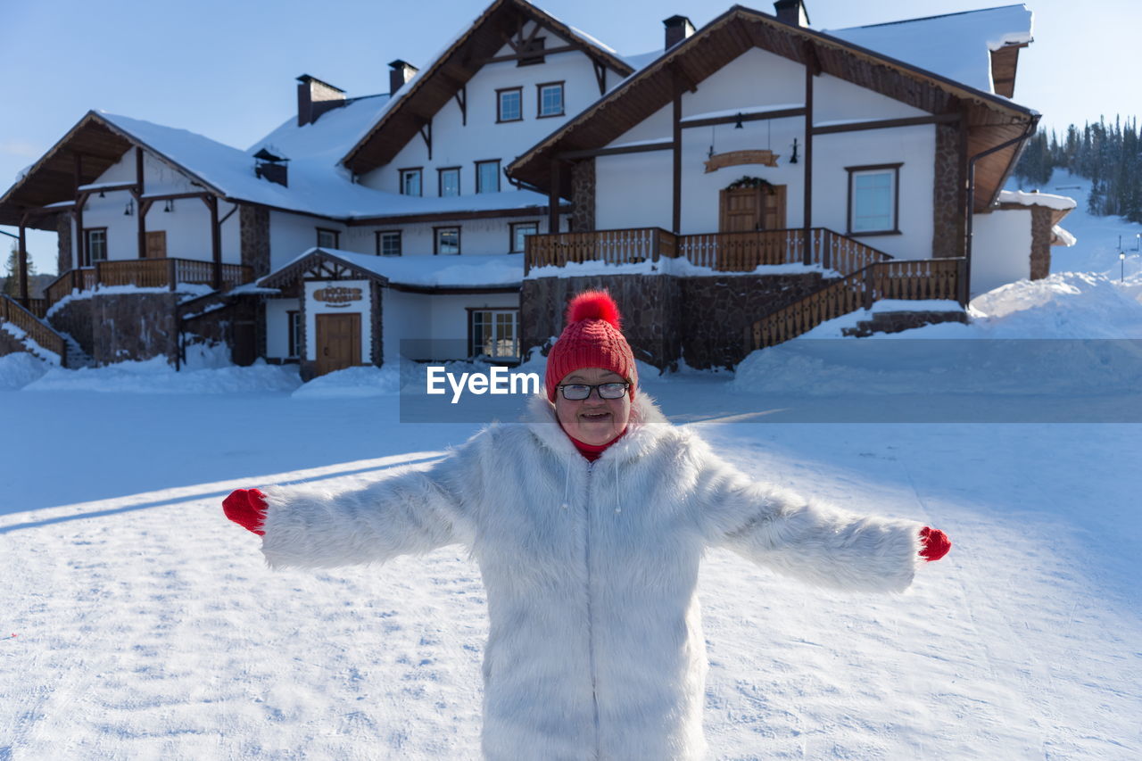 An elderly smiling woman stands with arms outstretched in front of the cottage on a sunny winter day