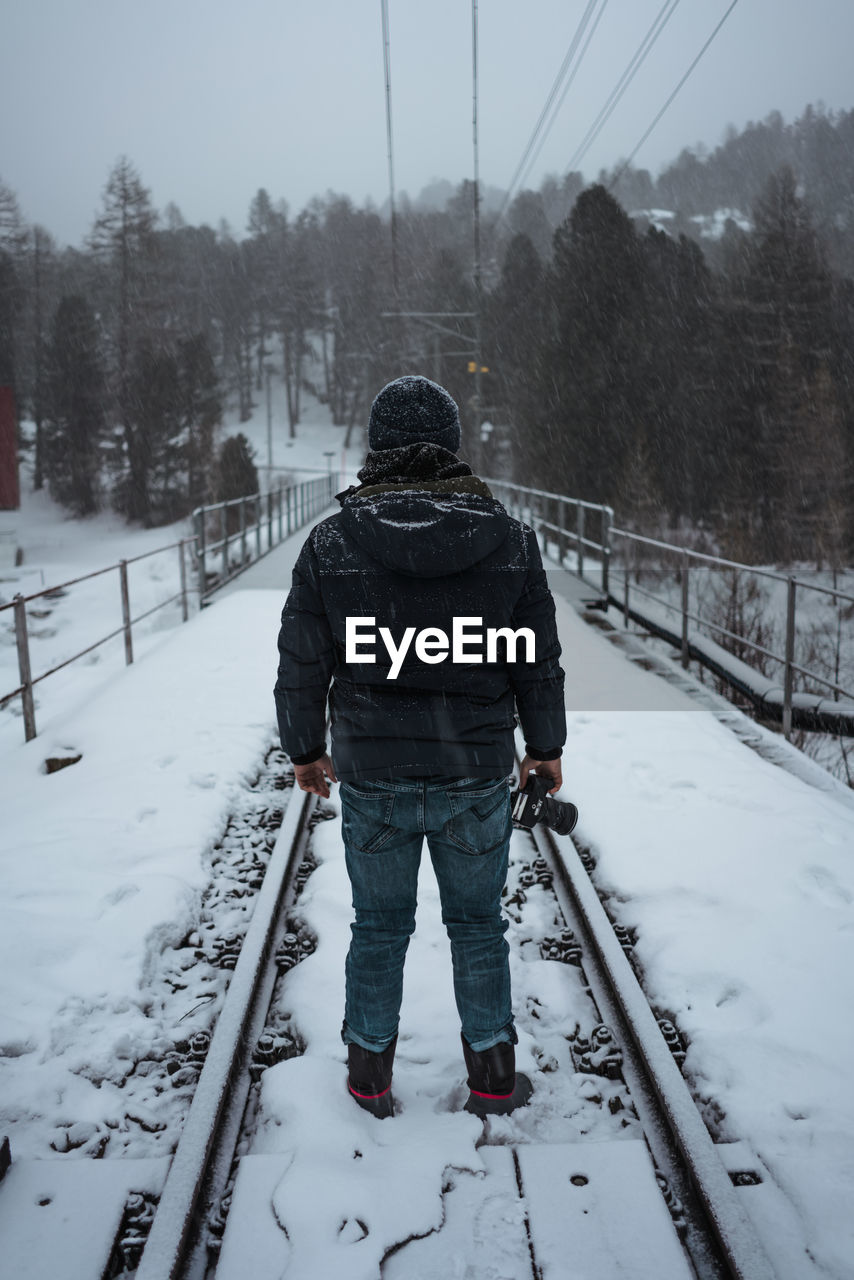 Rear view of young man standing on snow covered railroad track