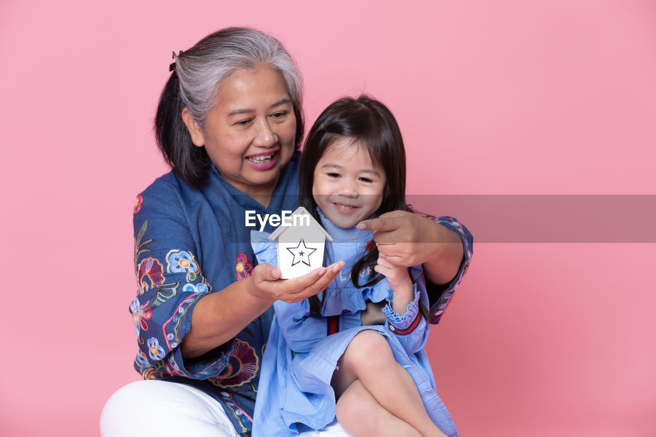 Smiling grandmother showing model home to granddaughter against pink background