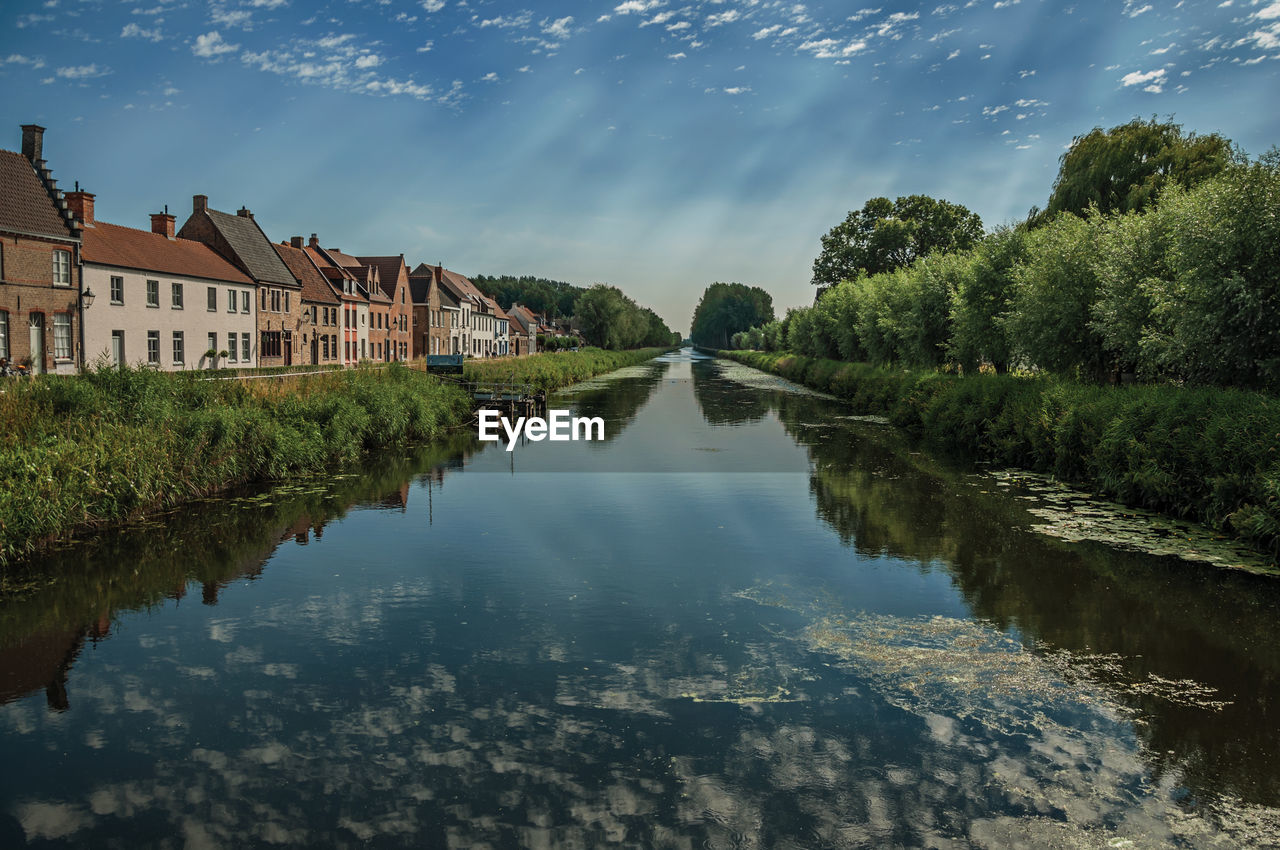 Trees and houses along canal with sky reflected on water in damme. a country village in belgium.