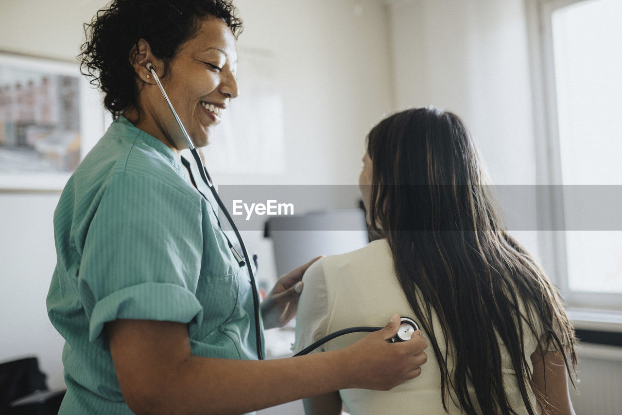 Side view of smiling mature female doctor examining young woman with stethoscope in clinic