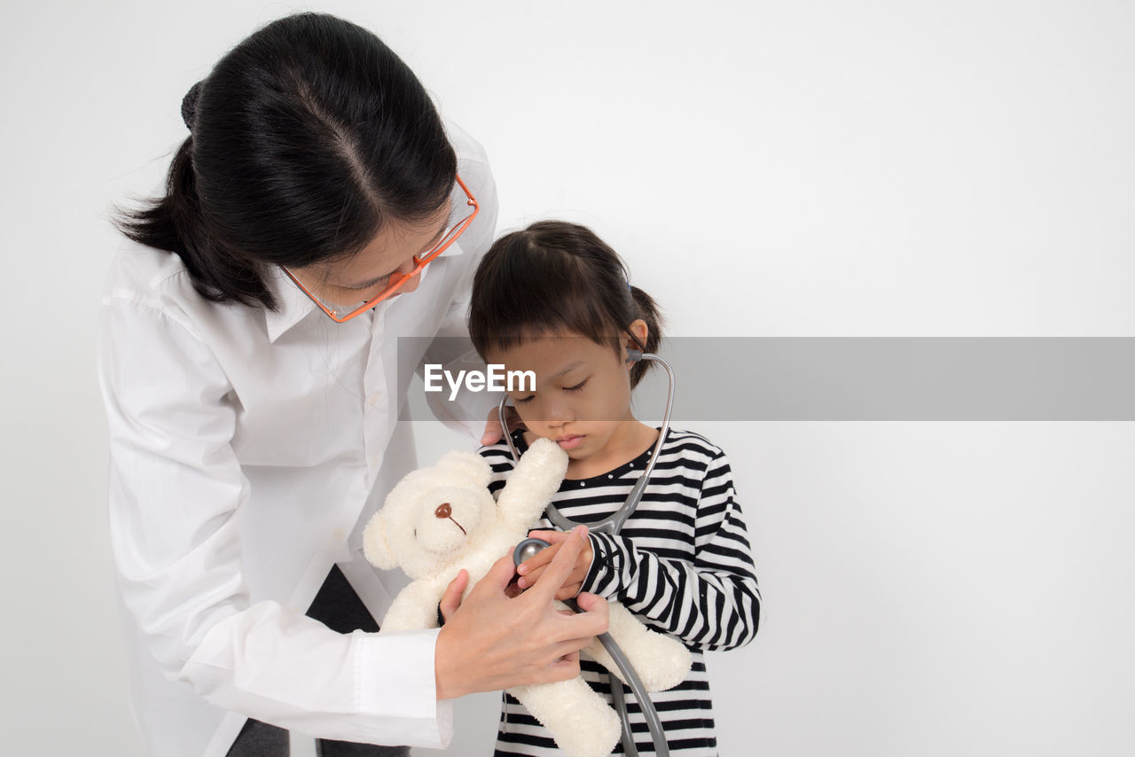 Doctor with girl holding stethoscope on teddy bear while standing against white background