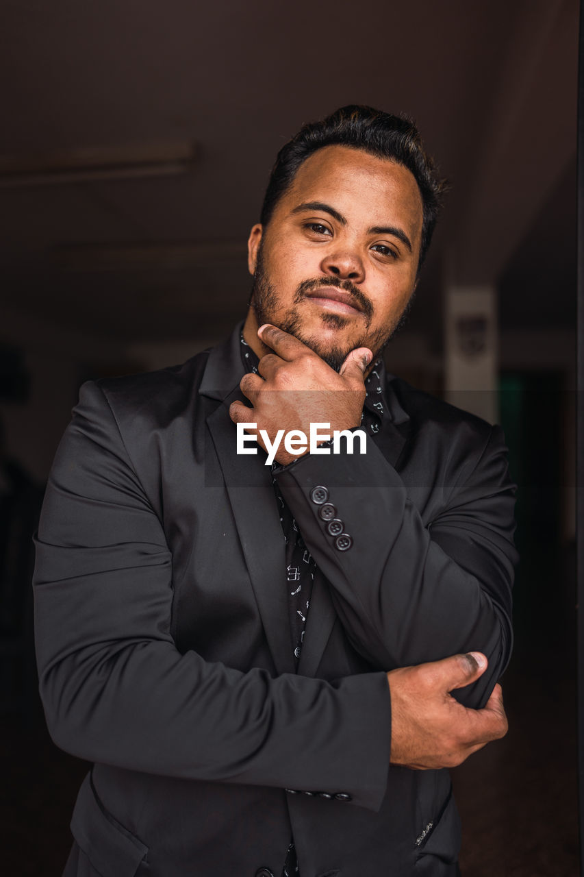 Self assured black male with beard in elegant suit standing with hand at chin and looking at camera