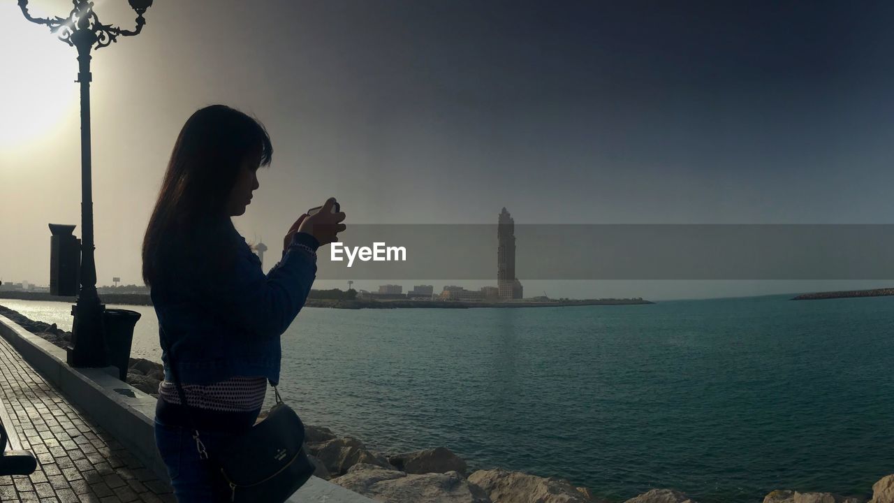 WOMAN PHOTOGRAPHING SEA AGAINST SKY