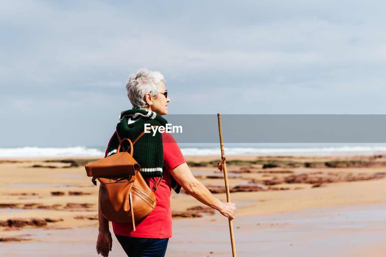 Side view of elderly female backpacker with trekking pole strolling on boulders against stormy ocean under cloudy sky