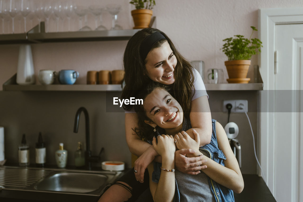 Portrait of happy woman embracing female friend from behind while sitting in kitchen at home