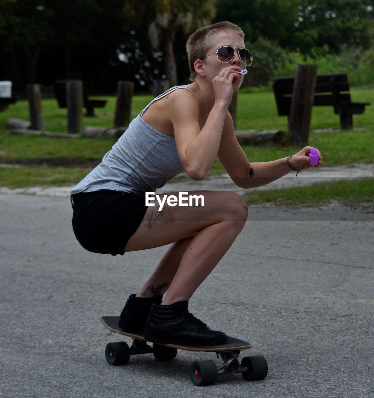 Full length of woman crouching on skateboard while blowing bubble over footpath