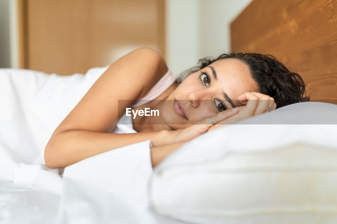 Calm female in pajama lying in bed on soft pillow under white blanket looking at camera