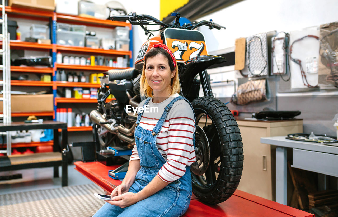 Mechanic woman holding phone sitting over platform with motorcycle on factory