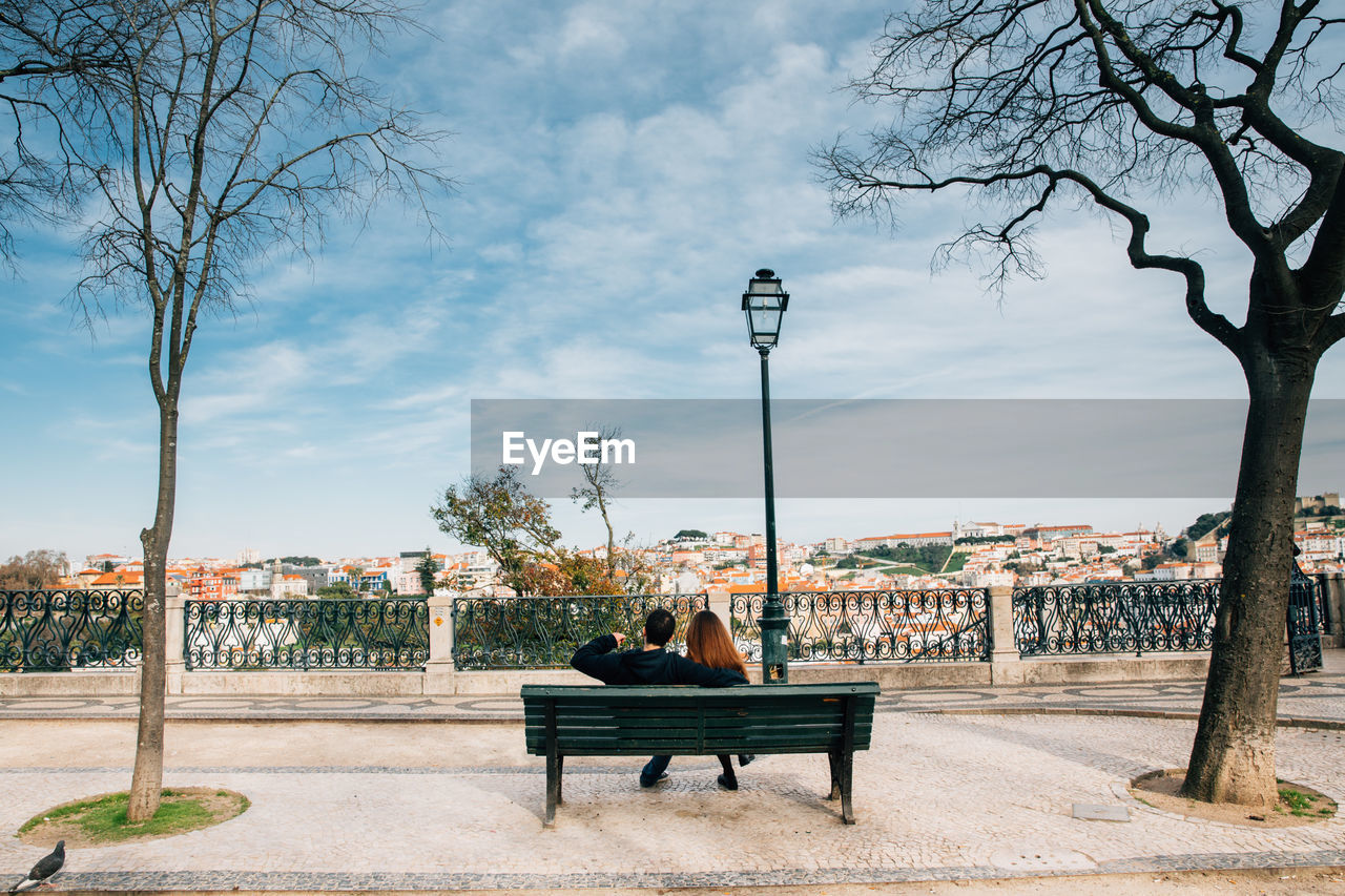 Rear view of couple sitting on bench in park against cloudy sky