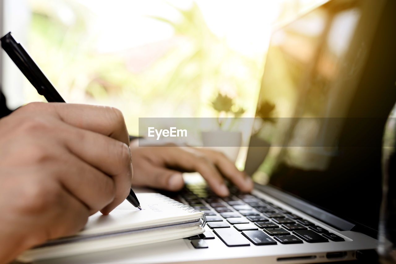 Cropped hands of man writing in book while using laptop on table