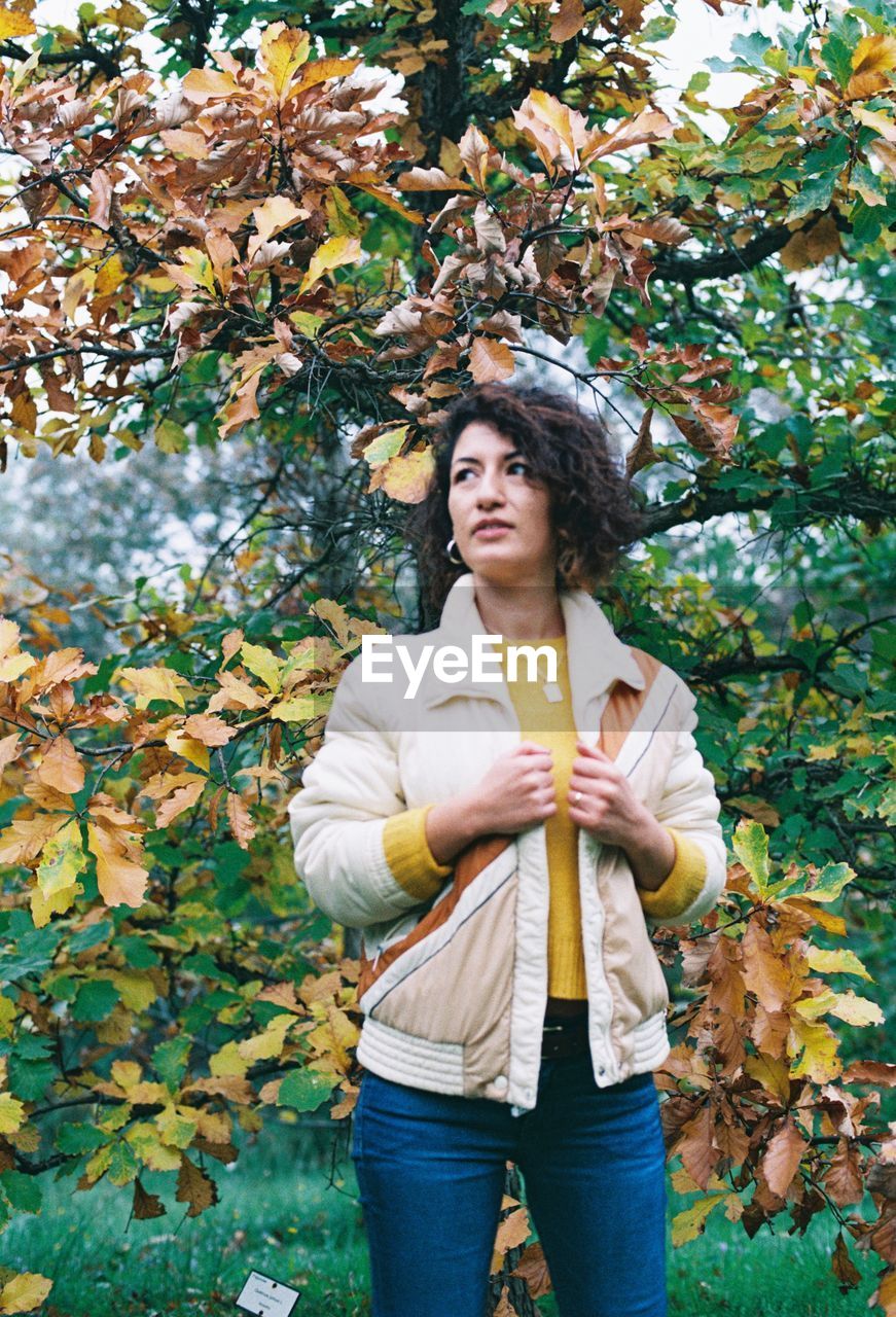 Woman in jacket looking away while standing against tree at forest