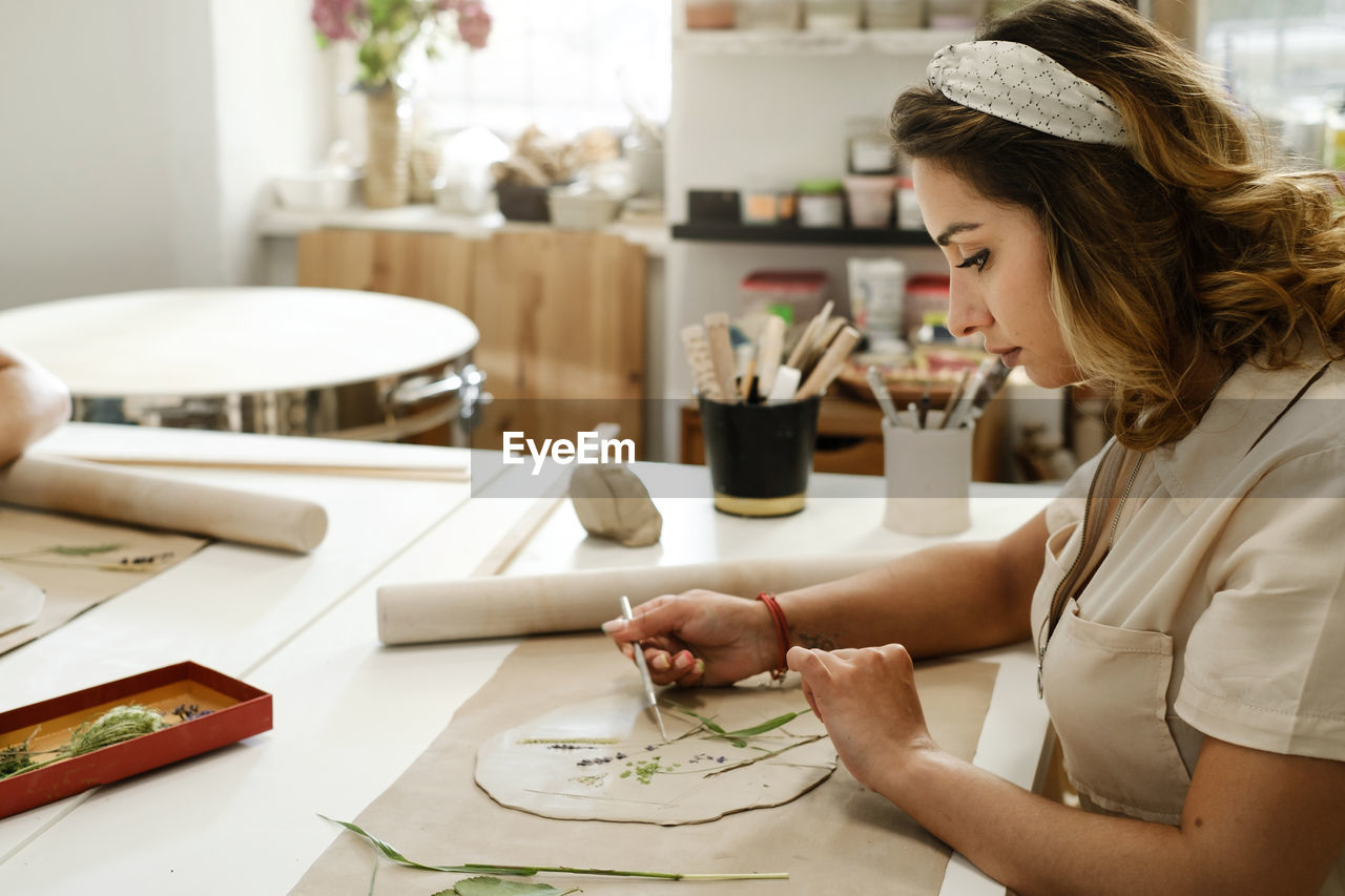 Woman decorating with flowers rolled clay, making ceramic plate in studio with floral pattern