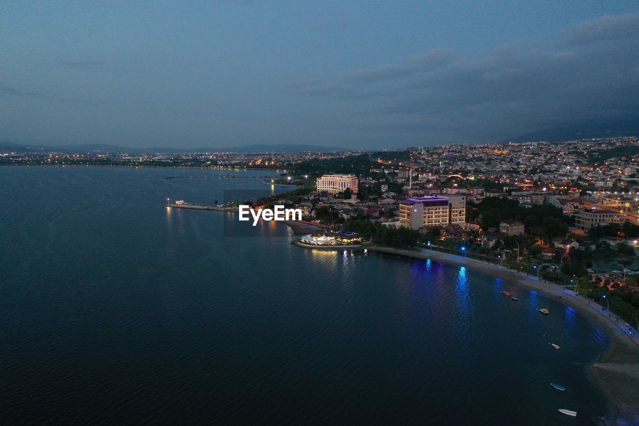 HIGH ANGLE VIEW OF ILLUMINATED CITY BY RIVER AGAINST SKY