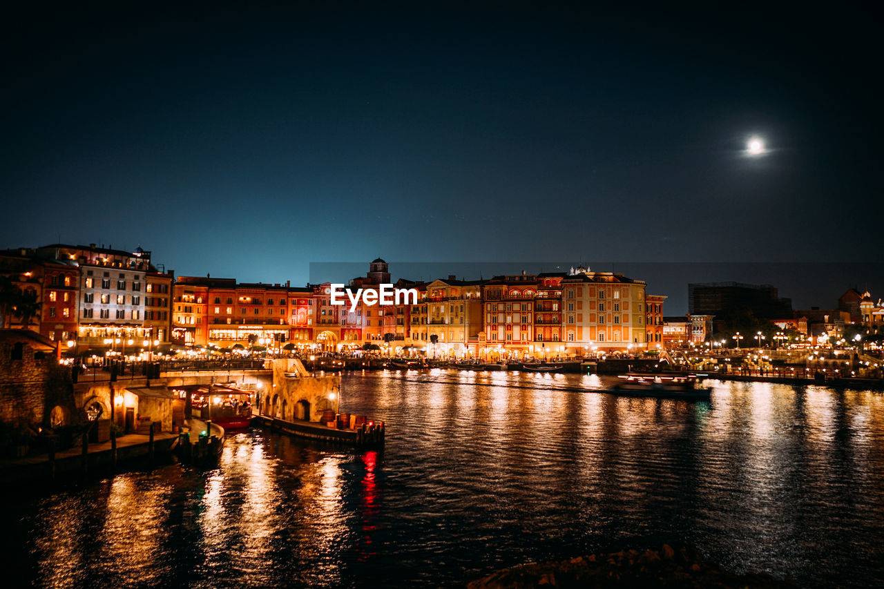 Illuminated buildings by river against sky at night