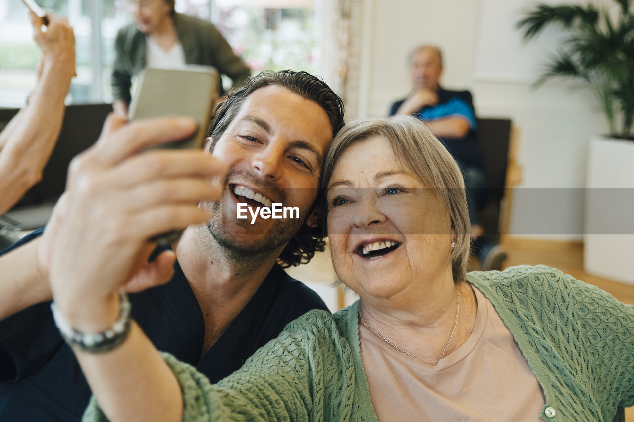 Smiling senior woman taking selfie with male healthcare worker while sitting at retirement home