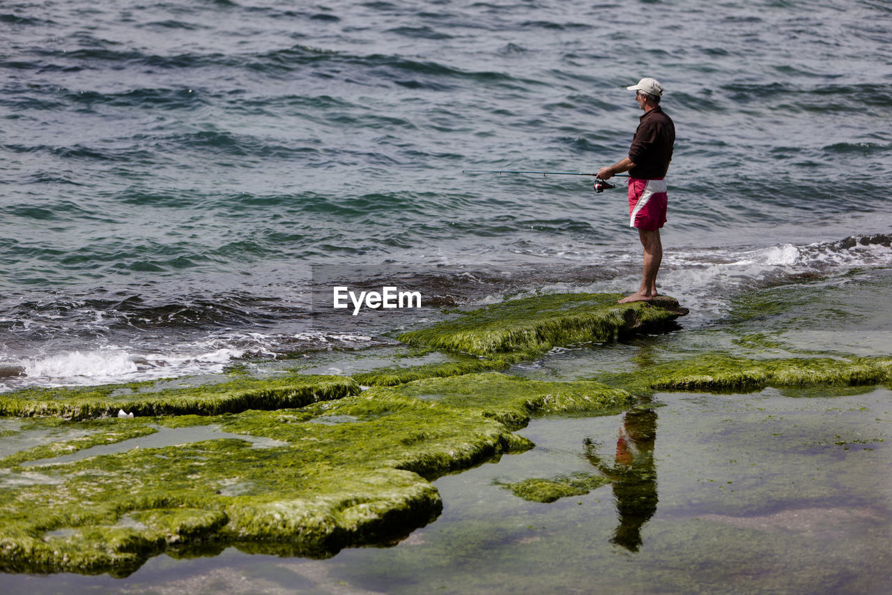 Mature man fishing while standing on moss covered rock by sea