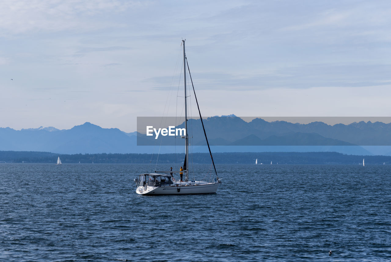 Sailboat off shore mid day sun with olympic mountains and people standing on deck.