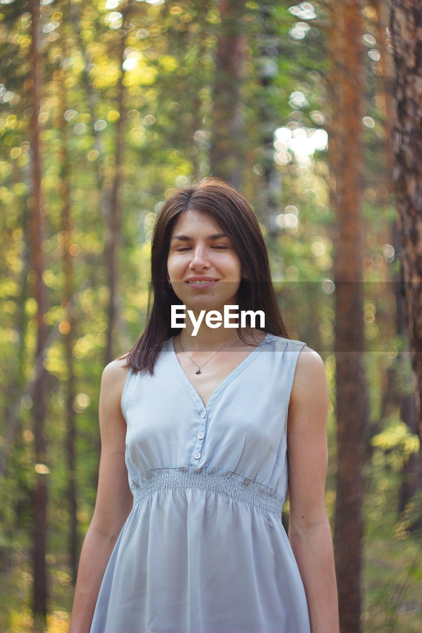 Smiling beautiful young woman with eyes closed in forest against trees