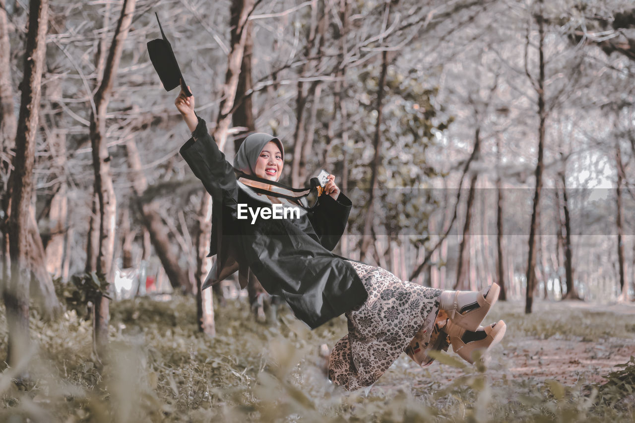 Full length of woman holding hat levitating against trees