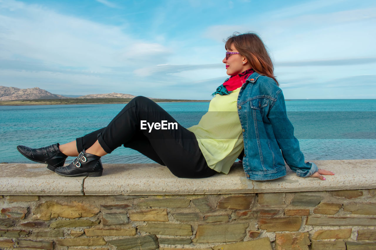 Side view of woman sitting on retaining wall against sea and sky