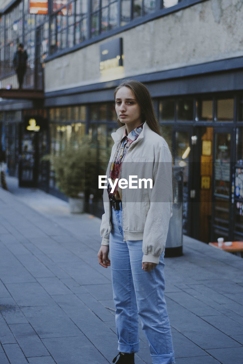 FULL LENGTH OF YOUNG WOMAN STANDING AGAINST CITY