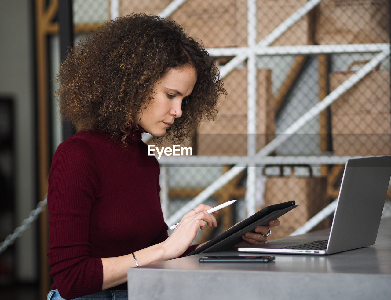 Beautiful woman with curly hair with pen in hand working with tablet while sitting at table in cafe.