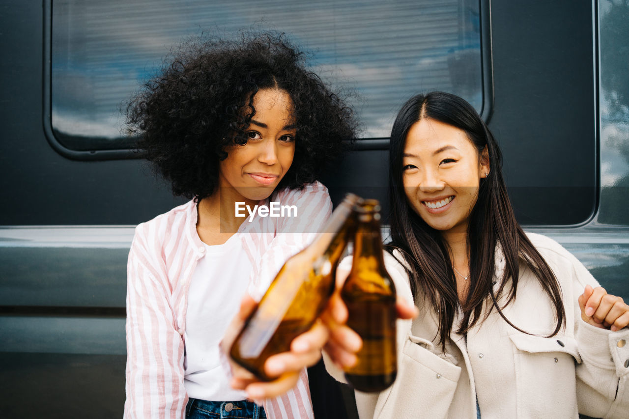 Happy young multiethnic female friends clinking bottles of beer while having fun and enjoying summer holidays together near camper van parked in nature