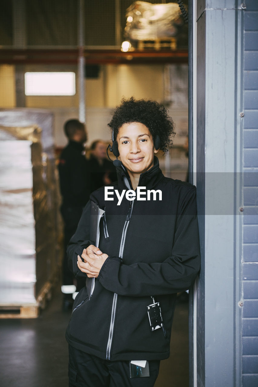 Confident female manual worker leaning on wall at logistics warehouse