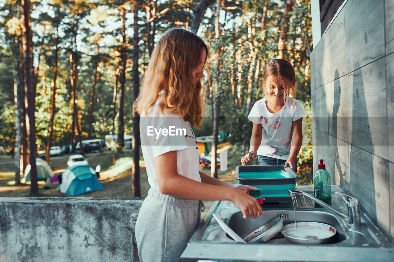 Teenager girl washing up the dishes pots and plates with help her younger sister